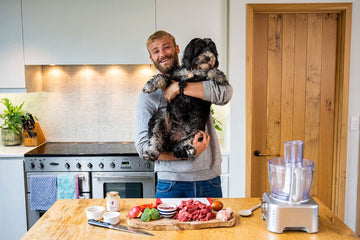 A man holding is dog in front of a tray of cut ingredients and a blender. 
